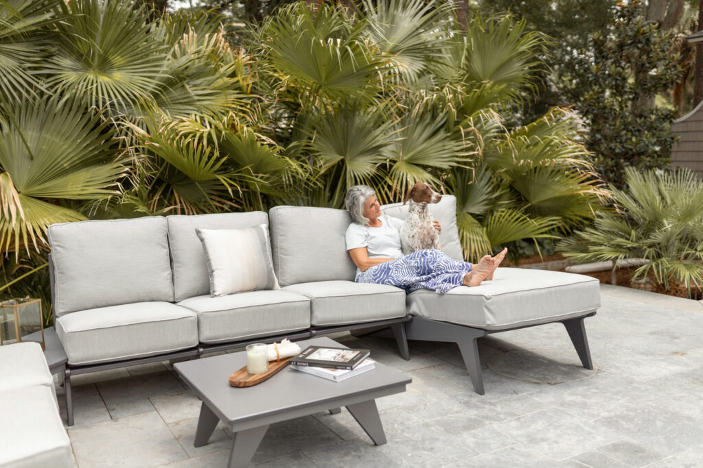A woman sitting on a gray outdoor sectional sofa, cuddling a dog, with lush palm trees in the background. A gray coffee table in front of the sofa holds a candle and a book.